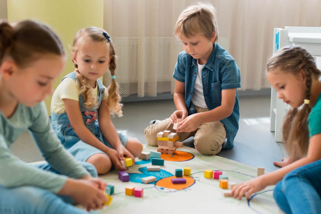 Zwei Kinder sitzen an einem Tisch und basteln konzentriert mit bunten Materialien unter Anleitung einer Erzieherin.