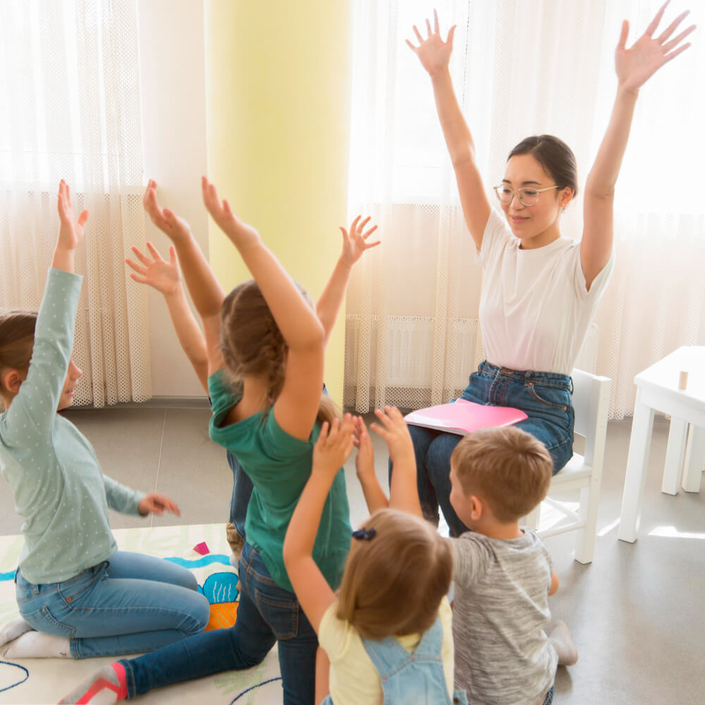 Eine Gruppe von Kindern sitzt im Kreis auf dem Boden und hört einer Erzieherin zu, die ihnen etwas erklärt.